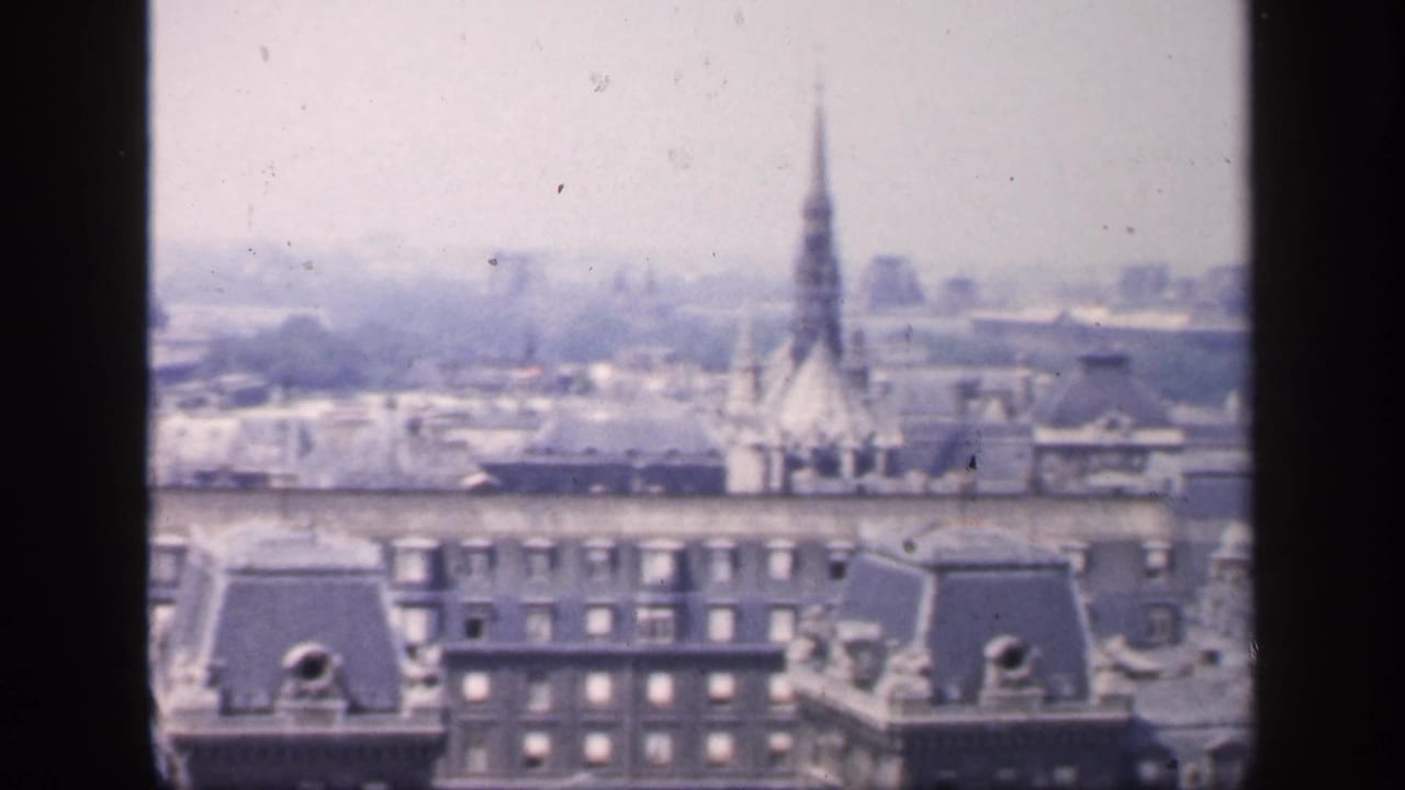 1949 France home-movie frame showing hazy Paris-like rooftops, dormers, chimneys and a distant spire above the skyline.