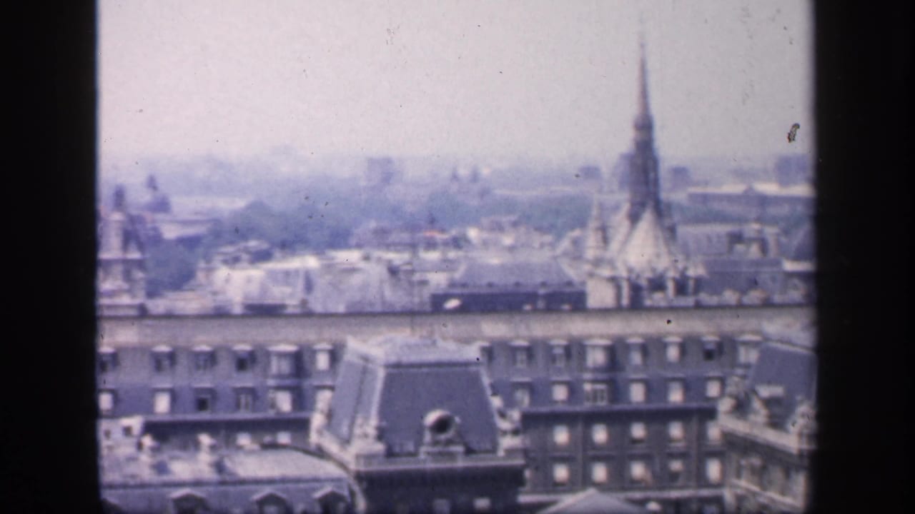 1949 France home-movie frame showing an elevated city view with mansard rooftops, dormer windows, and a tall church spire in haze.