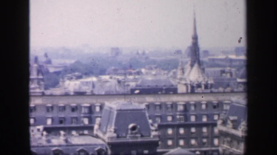 1949 France home-movie frame showing an elevated city view with mansard rooftops, dormer windows, and a tall church spire in haze.