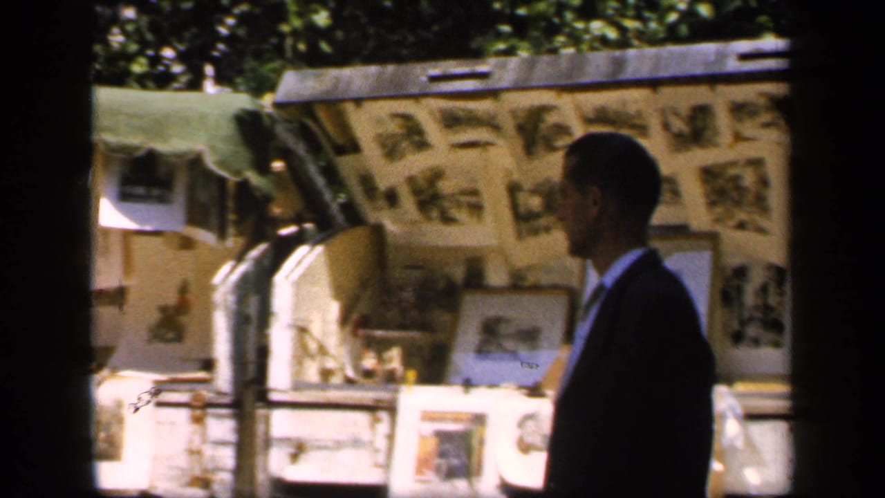 In 1949 France, a suited man passes sunlit outdoor stalls displaying pictures and photographs beneath a green canopy.