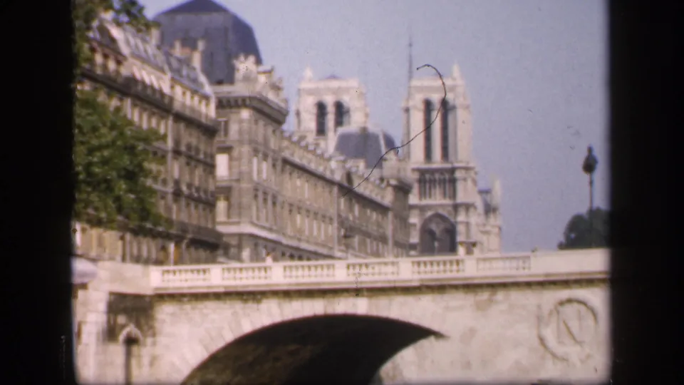 1949 Paris home-movie view of Notre-Dame Cathedral rising behind a stone bridge and adjoining riverside architecture.