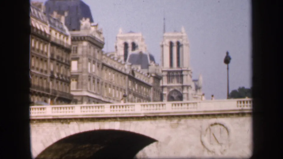 Visitor-shot 8mm frame from Paris in 1949 showing Notre-Dame's towers above riverside buildings and a stone bridge.