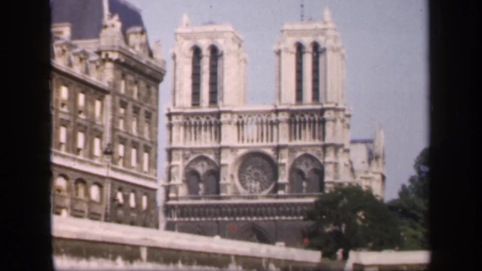 1949 8mm frame of Notre-Dame's facade from a bridge-level perspective, with a riverside building and parapet in view.