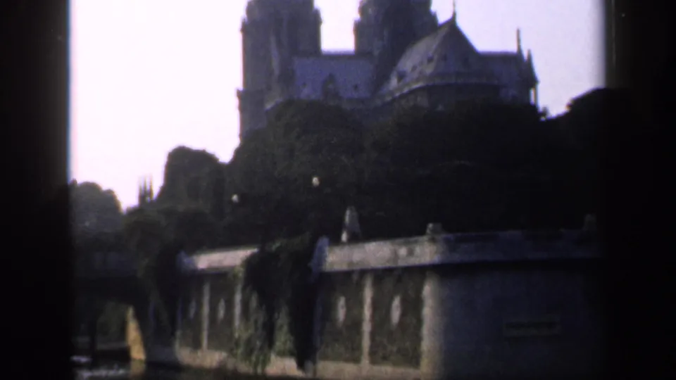 1949 Paris frame of Notre-Dame Cathedral backlit above trees, with the low wall and dark foreground giving the shot a shadowed home-movie look.