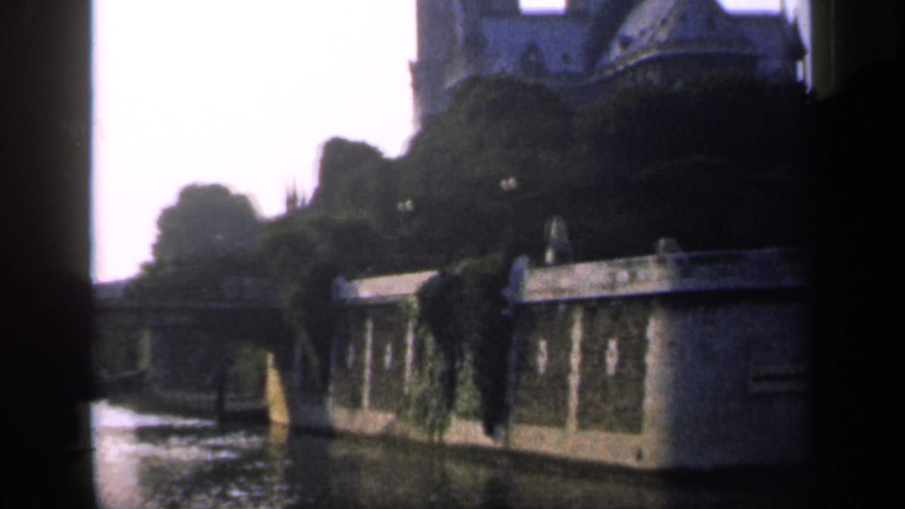 1949 France home-movie frame showing a river or canal beside stone walls, ivy, trees, and dark gate-edge obstruction.
