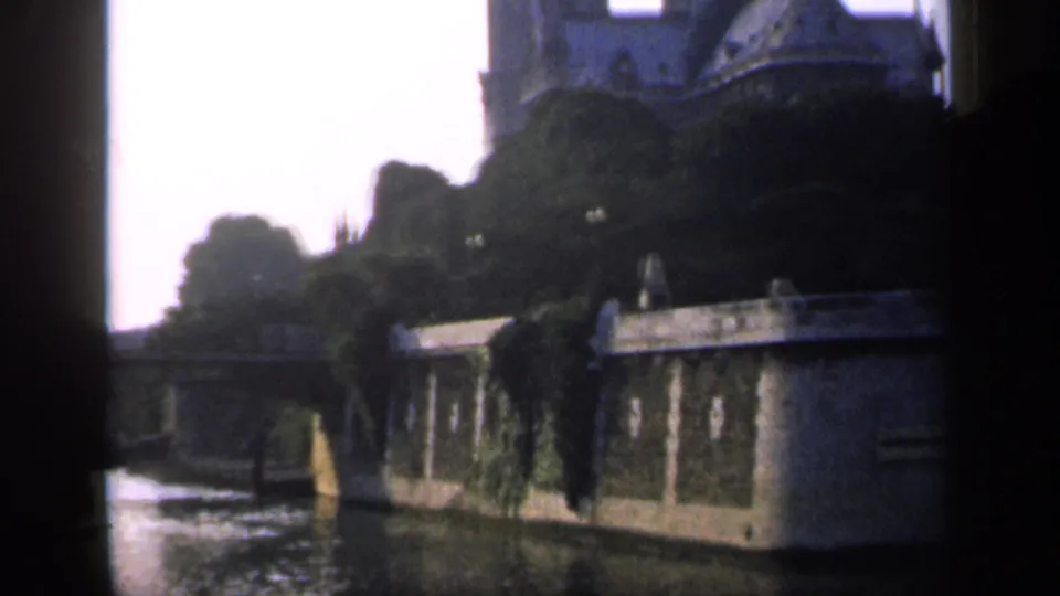 1949 France home-movie frame showing a river or canal beside stone walls, ivy, trees, and dark gate-edge obstruction.