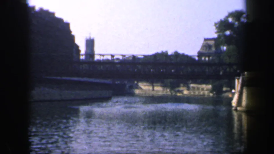 1949 France 8mm frame showing a river approach to a bridge, stone embankment, water reflections, and a small riverside structure.