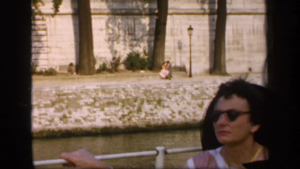 1949 France 8mm frame of a woman with short dark hair and sunglasses seated by a boat rail beside a stone riverside embankment.