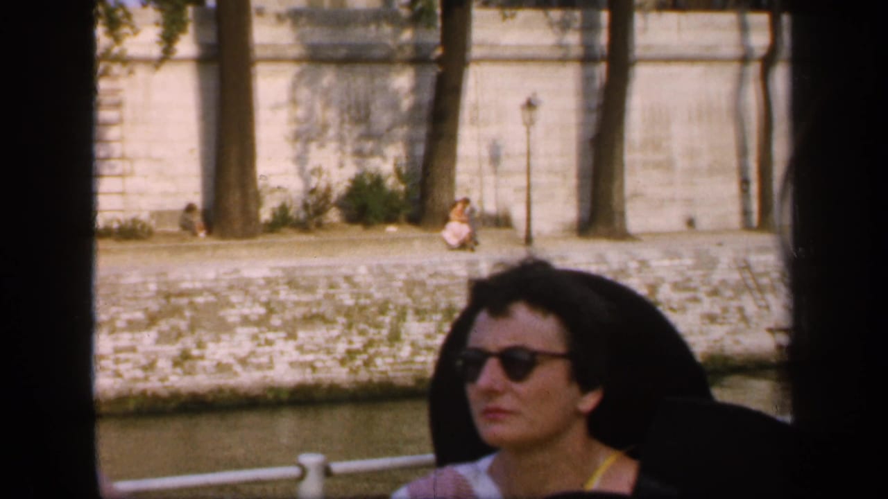 In 1949 France footage, a woman in sunglasses sits on a boat passing a stone canal embankment with trees and a lamppost behind her.