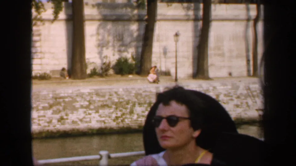 In 1949 France footage, a woman in sunglasses sits on a boat passing a stone canal embankment with trees and a lamppost behind her.