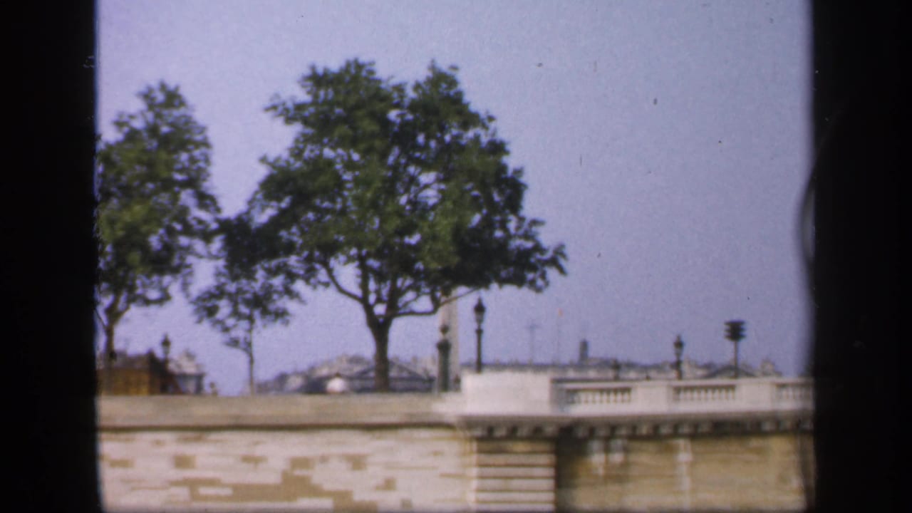 A boat passes a French waterside embankment with trees, railings, lampposts, and distant rooftops in a 1949 home-movie frame.