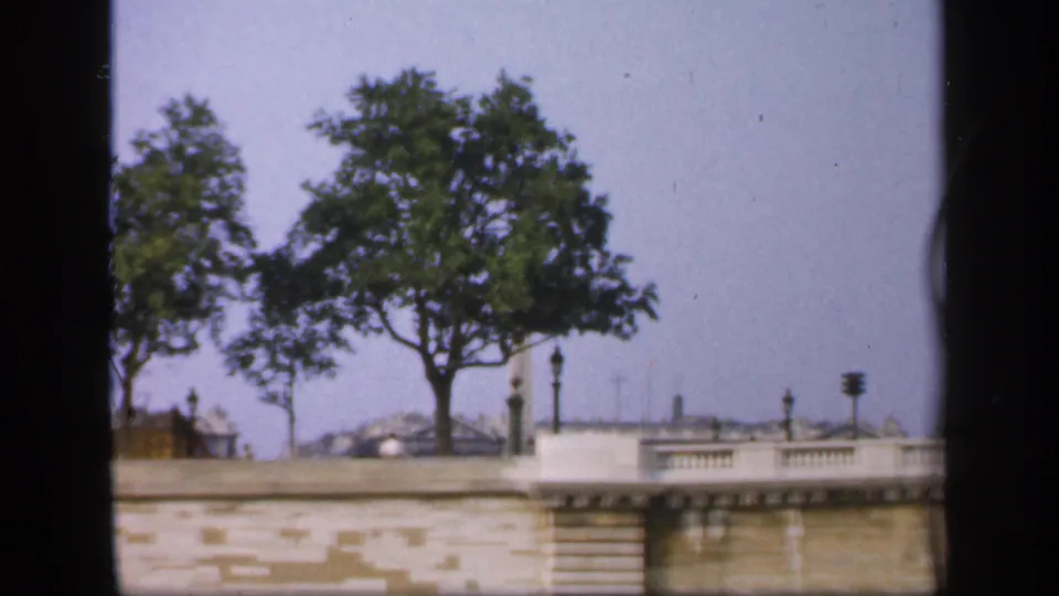 A boat passes a French waterside embankment with trees, railings, lampposts, and distant rooftops in a 1949 home-movie frame.
