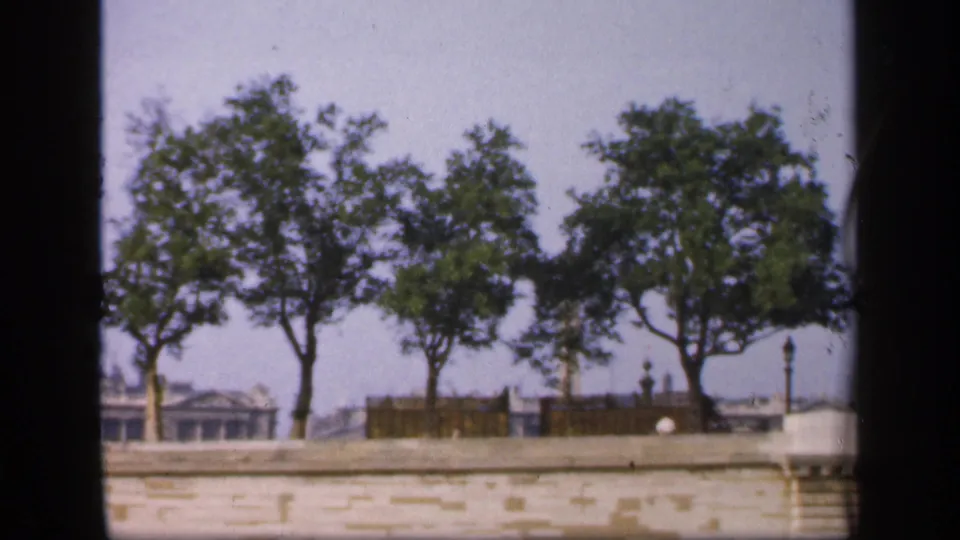 A 1949 France home-movie frame shows trees, railings, lampposts, and distant rooftops above a stone waterside embankment.