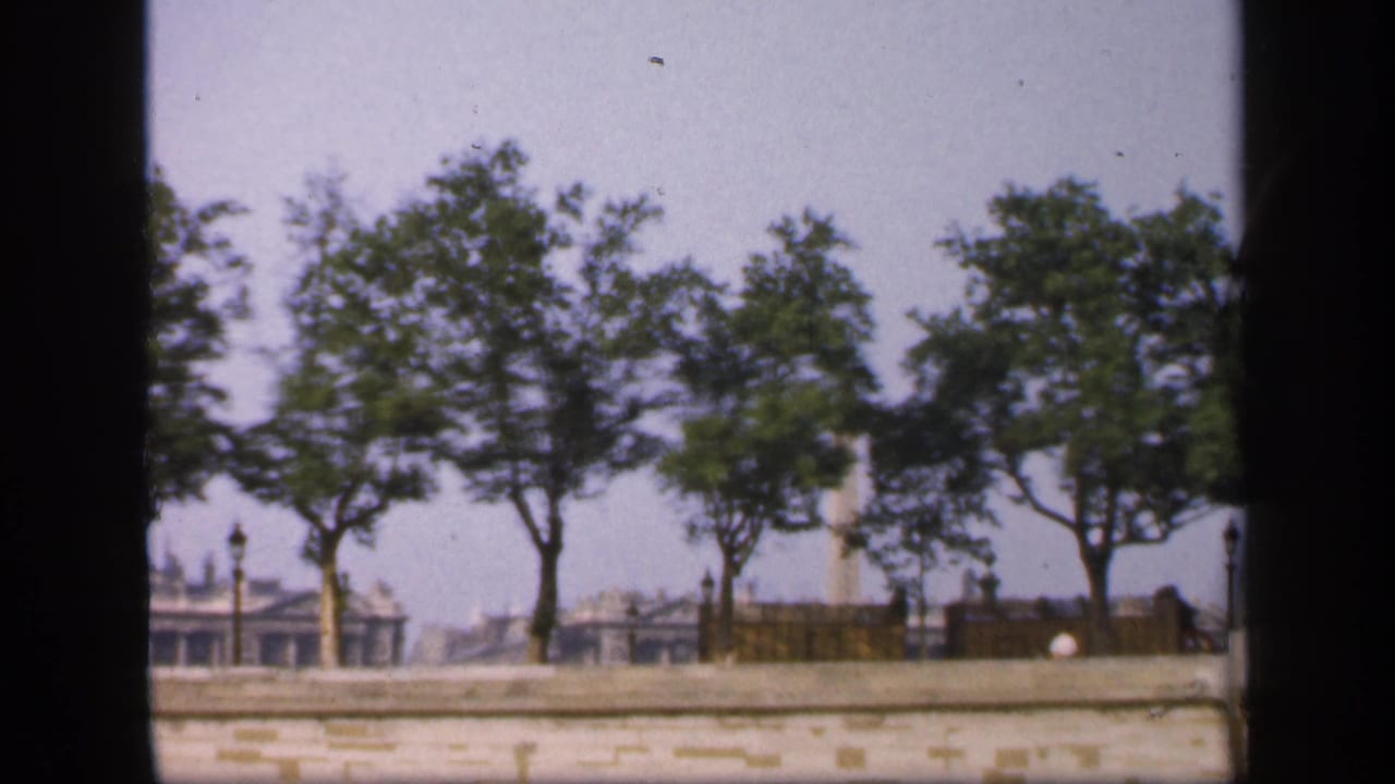 1949 France 8mm frame showing riverside trees, a pale embankment wall, lamp posts, and hazy distant buildings.