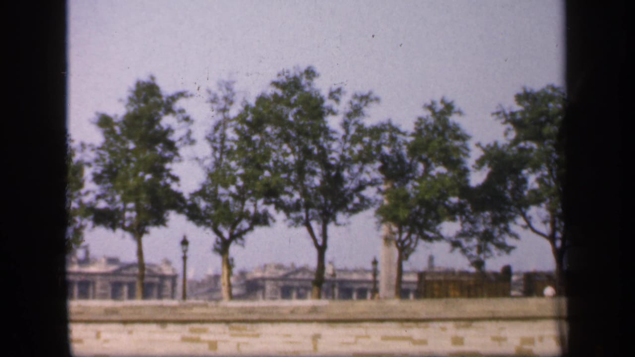 1949 France 8mm view of trees along a stone embankment, with low buildings and lamp posts softened by haze.
