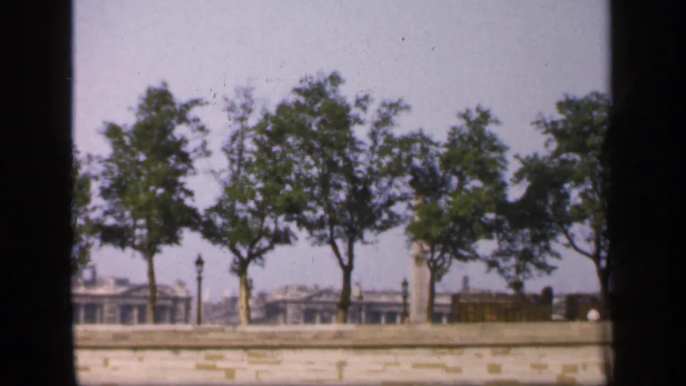 1949 France 8mm view of trees along a stone embankment, with low buildings and lamp posts softened by haze.