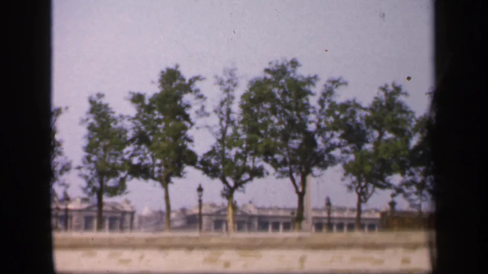 1949 France home-movie frame with clustered trees, a light stone wall, and distant city structures in soft focus.