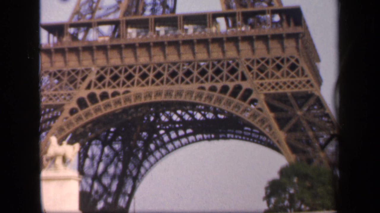 Lower Eiffel Tower structure seen from below with a pale sky and dark film border.