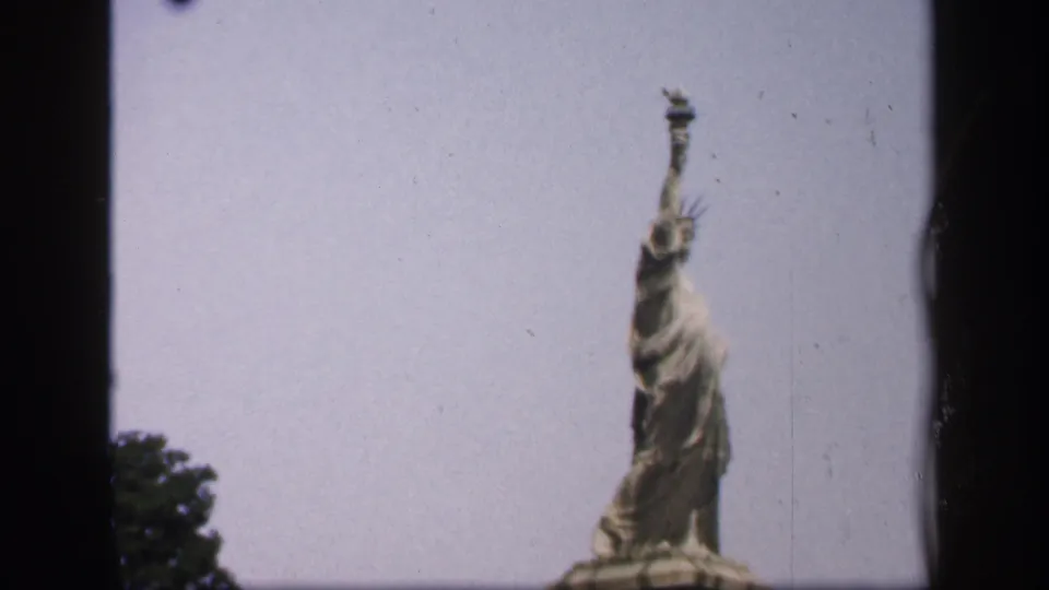 1949 New York 8mm frame showing the Statue of Liberty from a visitor viewpoint, with the torch raised into the pale sky and foliage at lower left.