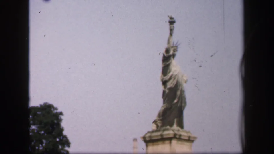 1949 8mm New York City home-movie frame showing a small Statue of Liberty replica on a pedestal beside a leafy tree.