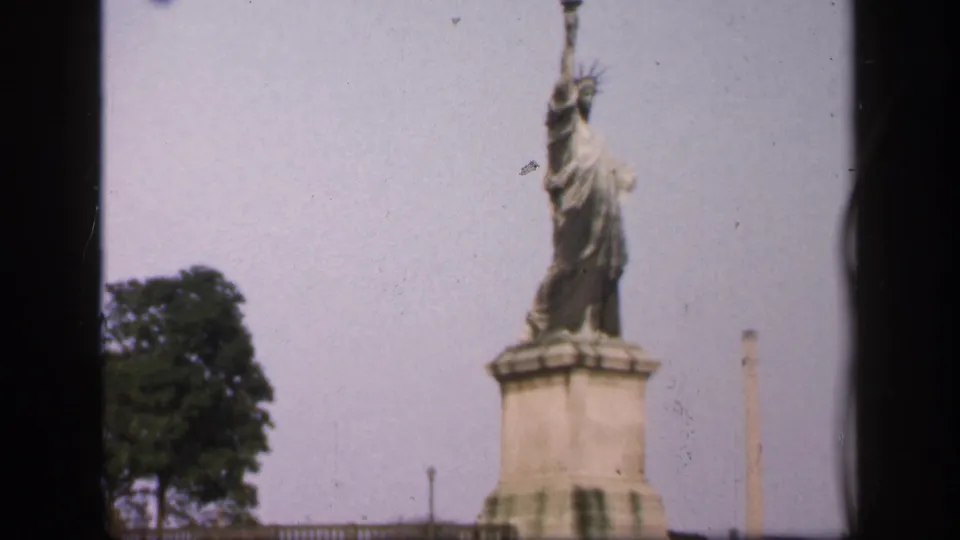 1949 New York City 8mm frame showing a small Liberty statue replica on a pedestal, with tree foliage and a vertical pole in view.