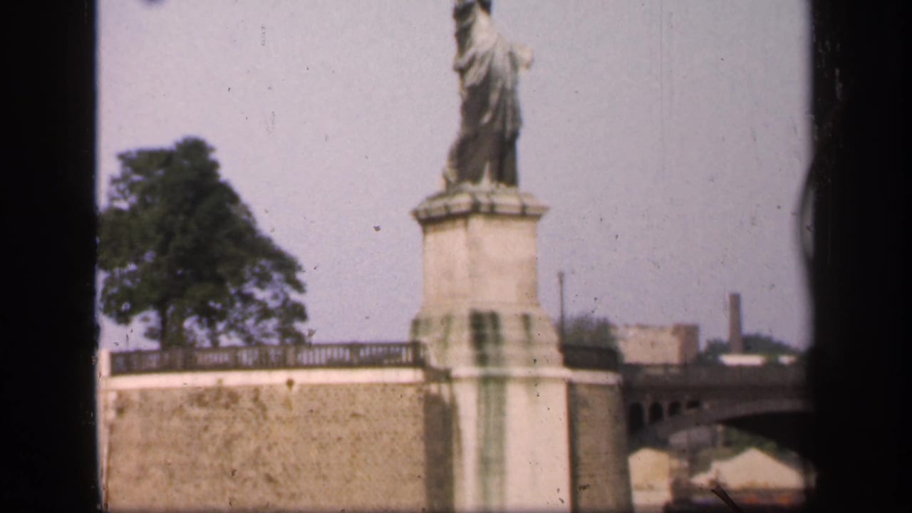 The replica monument appears above a stone embankment, with trees and distant industrial-looking structures in the background.