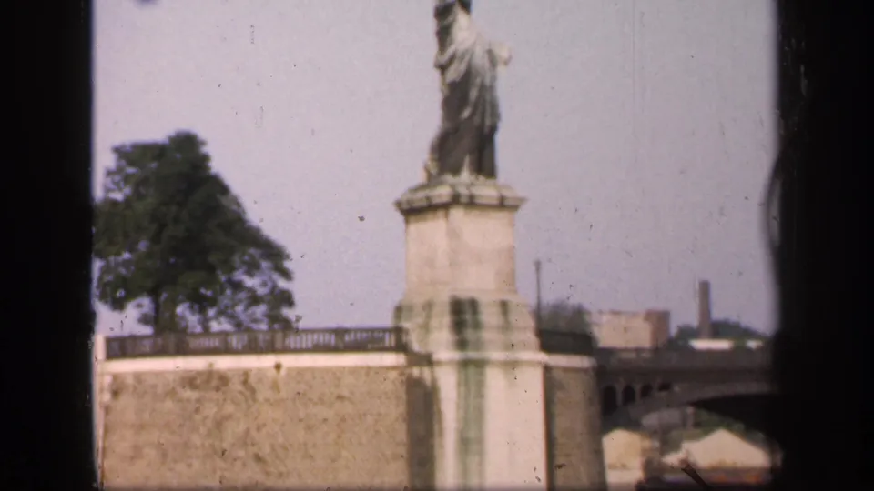 The replica monument appears above a stone embankment, with trees and distant industrial-looking structures in the background.