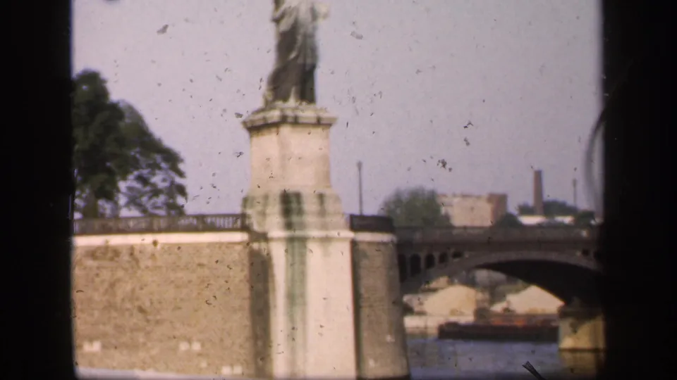 1949 home-movie frame showing the statue pedestal, stone embankment, and an arched bridge crossing the river to the right.