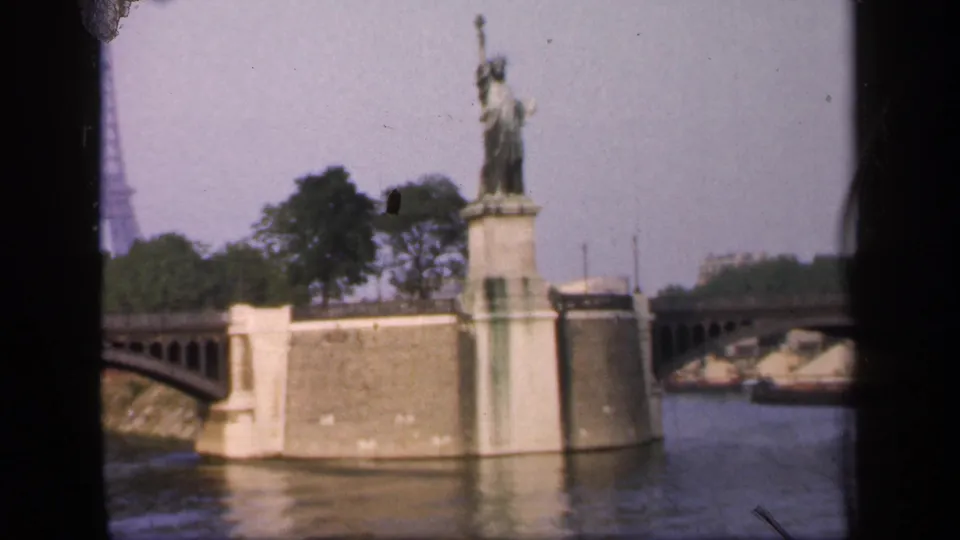 Wider 1949 France view of the Statue of Liberty replica by the river, with a bridge, trees, water, and the Eiffel Tower faintly in the distance.