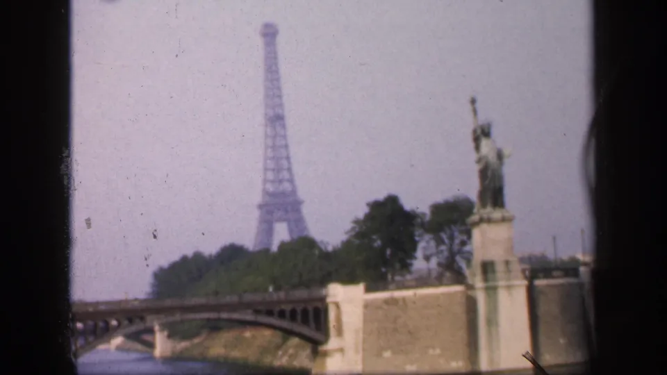 Soft 1949 Paris home-movie frame showing the Eiffel Tower behind trees, a bridge, and the Statue of Liberty replica near the Seine.