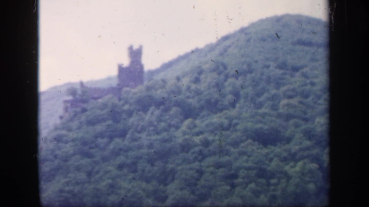 A soft 1949 Ireland landscape frame with dense green hillside, pale sky, and a distant castle-like tower near the ridge.