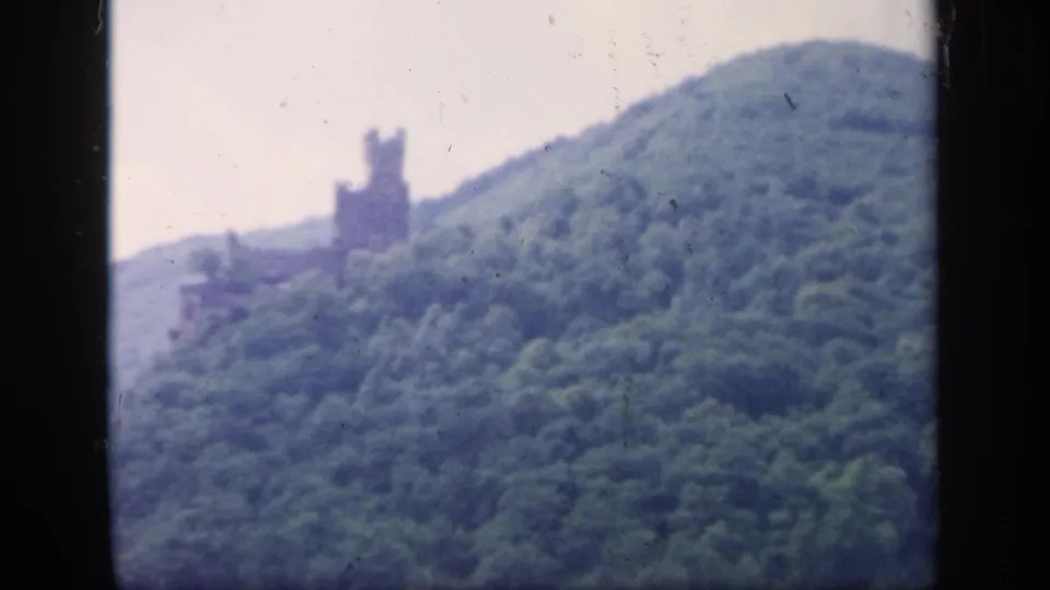 A soft 1949 Ireland landscape frame with dense green hillside, pale sky, and a distant castle-like tower near the ridge.