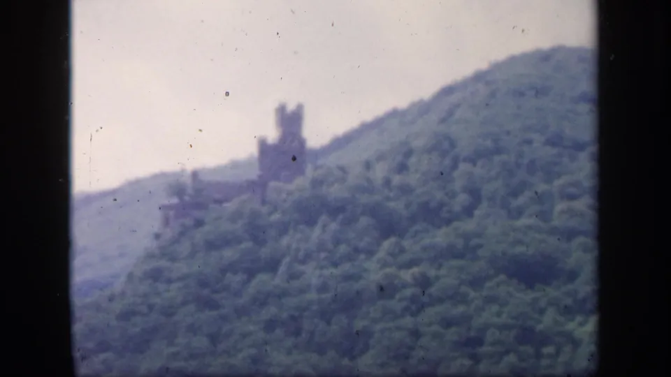 1949 Ireland home-movie frame showing a wooded slope, pale sky, and a distant stone tower or ruin softened by haze and film grain.