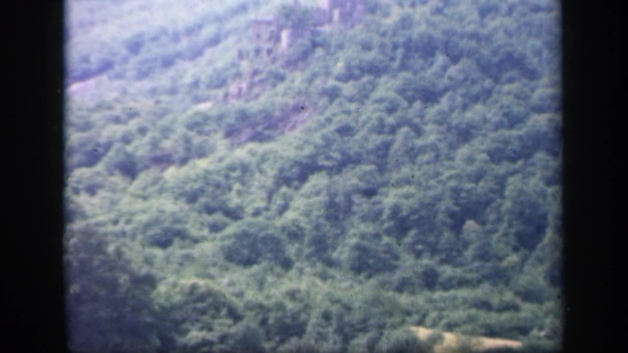 1949 Ireland 8mm frame showing a green wooded hillside and a distant grey stone ruin under a pale cloudy sky.