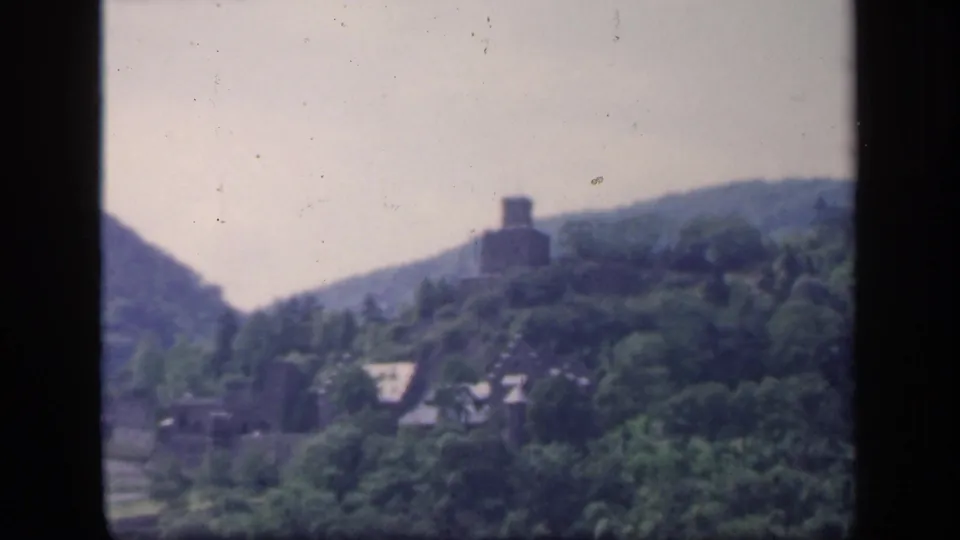 1949 Ireland 8mm frame with a castle-like ruin on a wooded hill, rooftops below, and soft valley scenery.