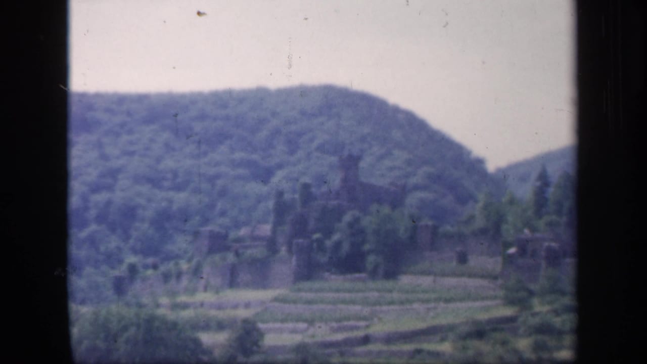 Soft distant view of castle ruins below wooded hills with terraced fields in front.