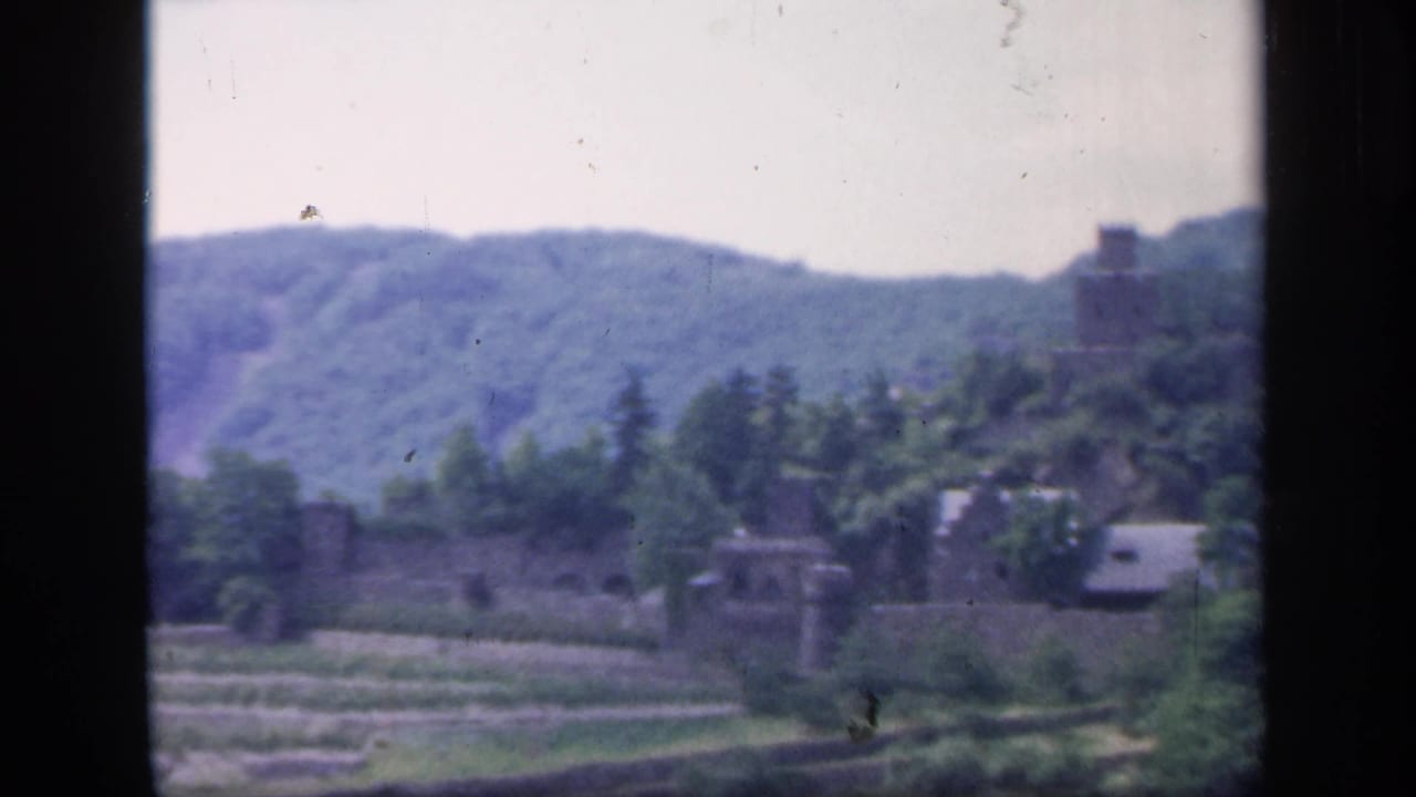 Distant castle ruins beyond terraced fields and trees with wooded hills behind.