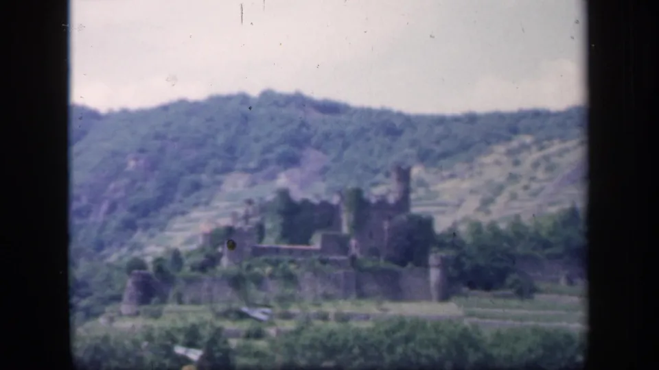 Vintage 8mm frame from Ireland showing the castle lower in frame, with rolling hills, shrubs, terraces, and a cloudy sky.