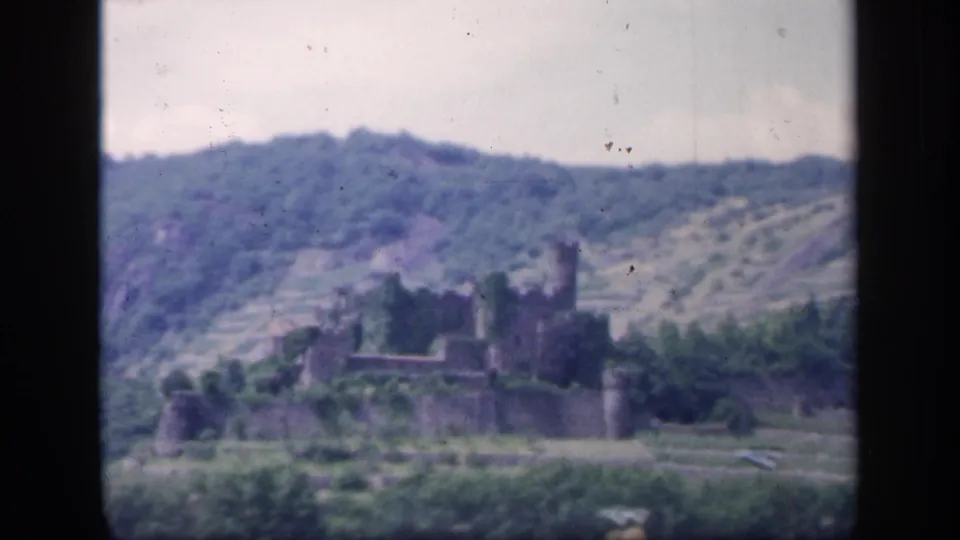 1949 Ireland 8mm frame with a distant stone castle, green terraced slopes, wooded hill backdrop, and visible film grain.