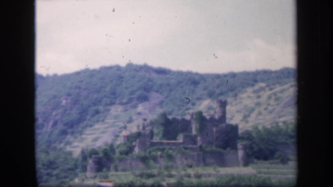 Distant castle ruin below a wooded ridge, framed by black film borders.