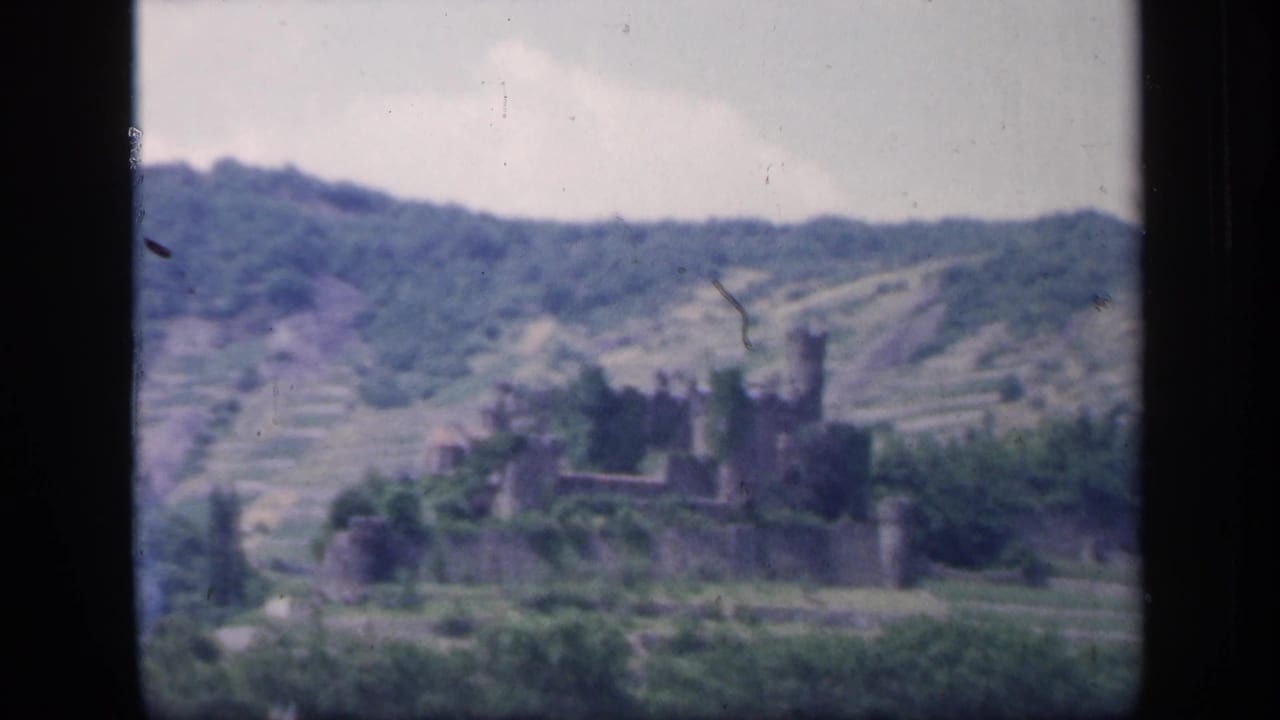 Distant castle ruin on a green hillside with terraced slopes and dark film-frame edges.