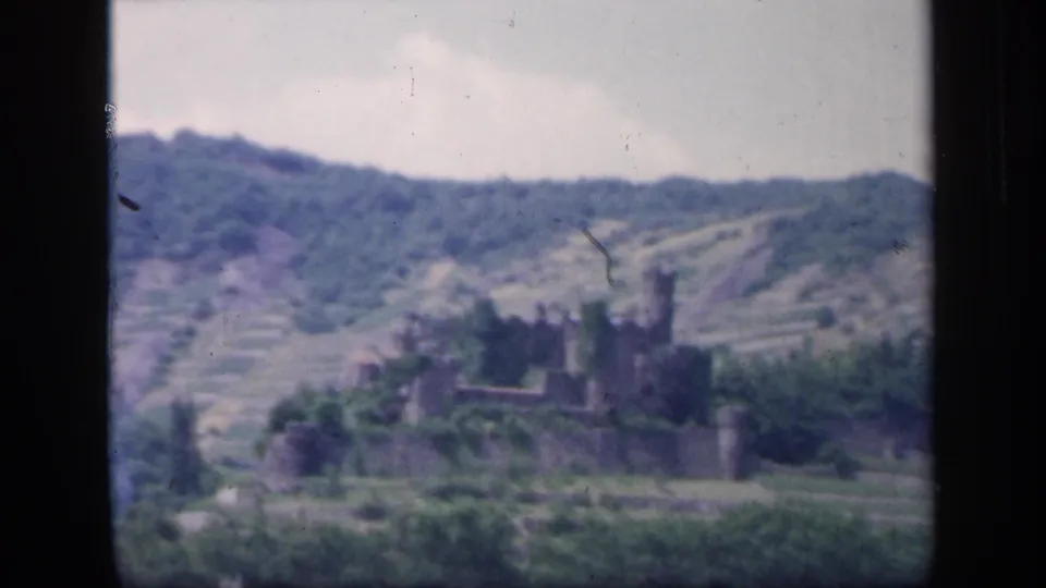 1949 Ireland 8mm frame showing a distant stone castle ruin among trees on a terraced hillside under a pale sky.