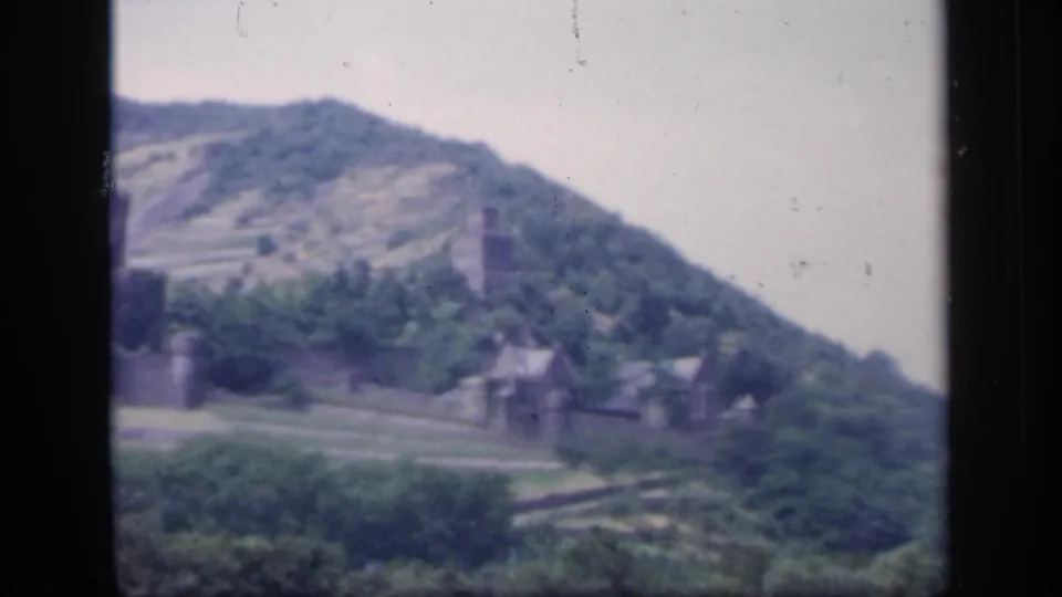 1949 Ireland 8mm frame with a stone tower, hillside buildings, trees, and terraced slopes in soft focus.