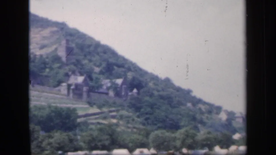 1949 Ireland home-movie frame showing a hillside ruin, small buildings, trees, and sloping terrain through dark gate edges.
