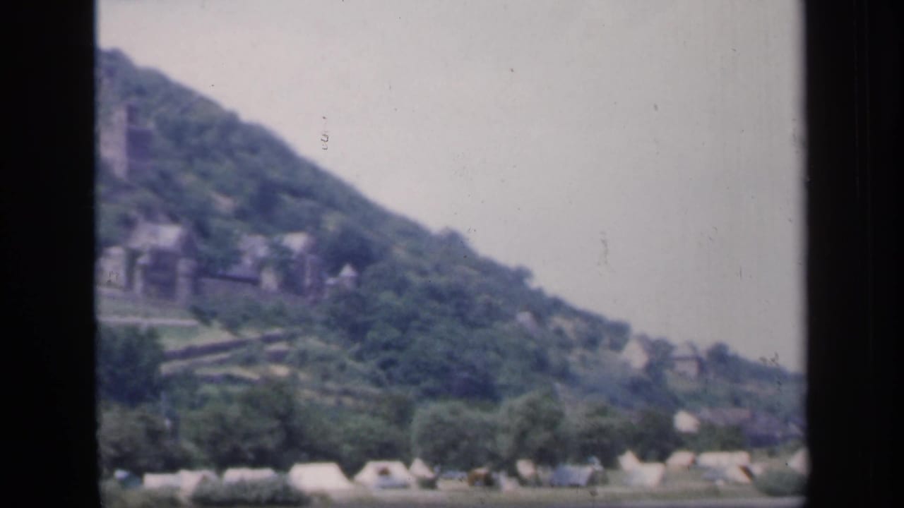 Tents along a river below a wooded hillside with distant buildings.
