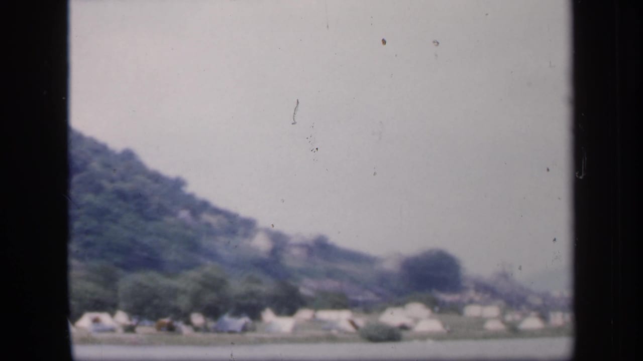 Campsite tents stretch along the water with green hills behind.