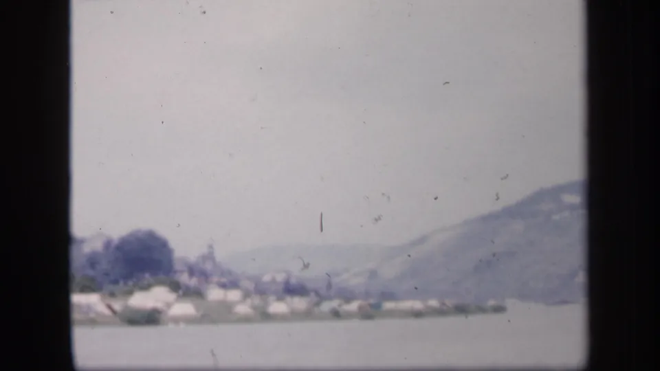 1949 Ireland 8mm frame showing a distant settlement or shoreline structures across water below a misty mountain.