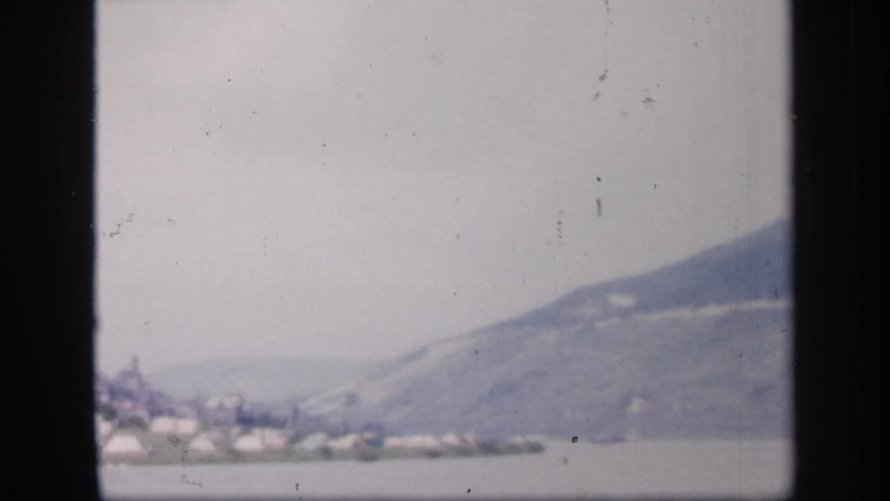1949 Ireland home-movie frame with small pale shoreline forms and a broad, mist-softened mountain beyond the water.