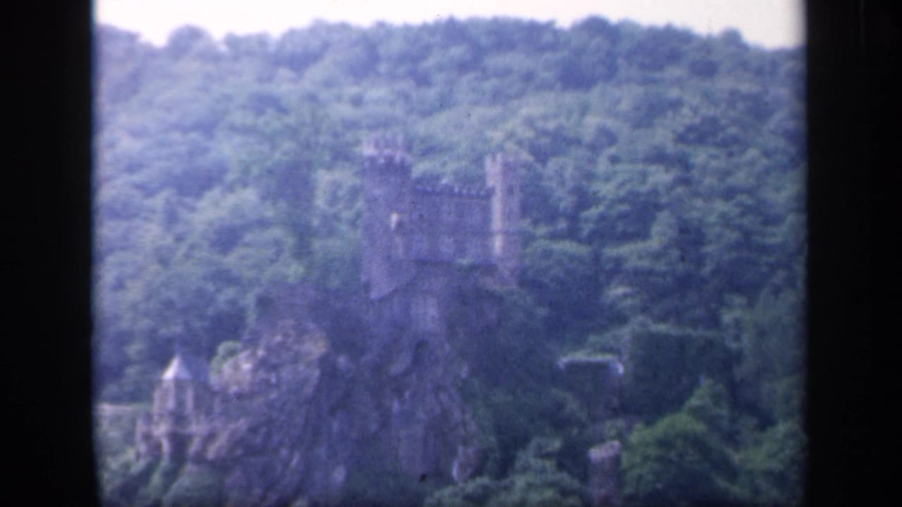 Distant stone castle on a rocky wooded hillside, framed by black 8mm borders.