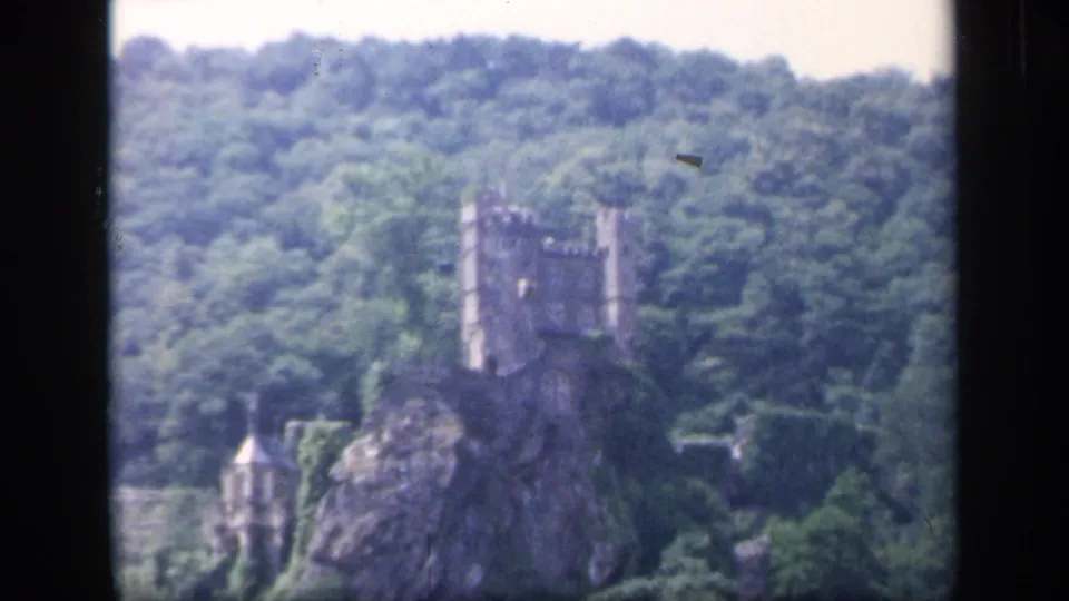 1949 Ireland home-movie frame with a distant castle above a rocky hillside, enclosed by woodland and visible 8mm frame edges.